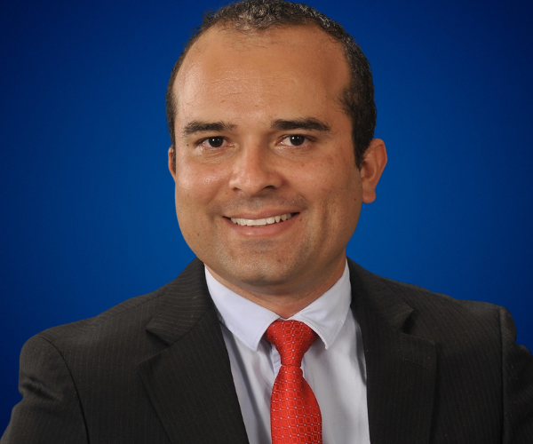 Sergio Rey sonriendo, conferencista internacional y experto en liderazgo y desarrollo organizacional, posando con traje formal sobre fondo azul.