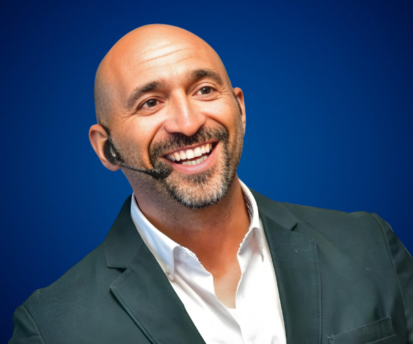 Sergio de la Calle sonriente en una conferencia, vistiendo saco oscuro y auricular, sobre fondo azul.