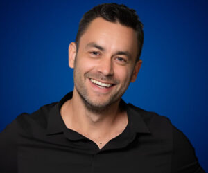 Fotografía de Achi Kushnir sonriendo, con camisa negra y fondo azul, transmitiendo cercanía y profesionalismo.