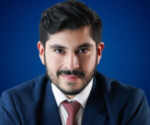 Fotografía de Edgar Dojaque con traje azul, camisa blanca y corbata roja, posando frente a un fondo azul, transmitiendo seguridad y profesionalismo.