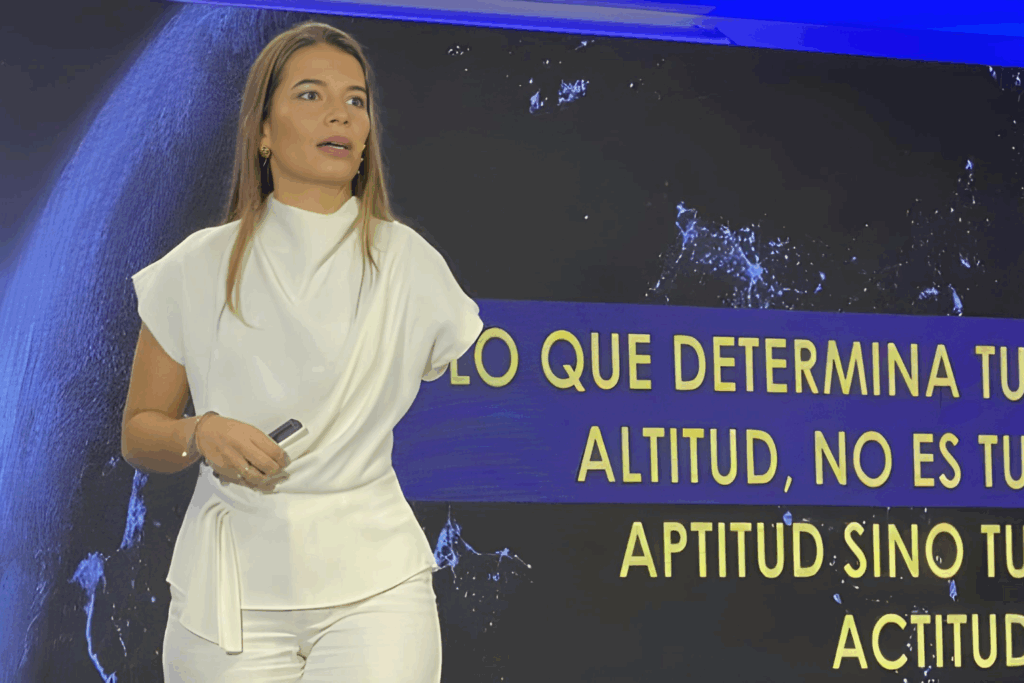 Fotografía de Emily Clavel impartiendo una conferencia, vestida de blanco, frente a una pantalla con la frase “Lo que determina tu altitud no es tu aptitud sino tu actitud”.