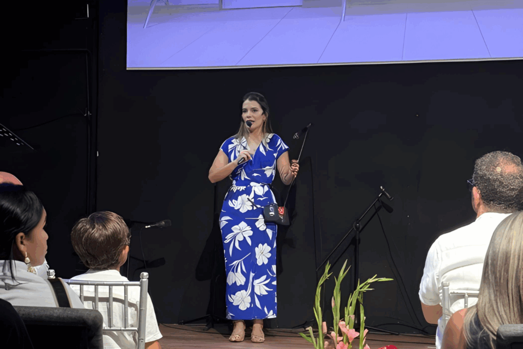 Fotografía de Emily Clavel impartiendo una conferencia, vestida con un elegante vestido azul con estampado floral blanco, dirigiéndose a una audiencia atenta.