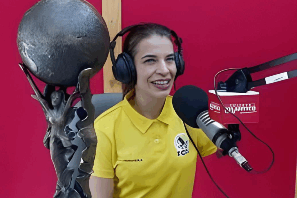 Fotografía de Emily Clavel en una cabina de radio, sonriendo con audífonos y micrófono profesional, vistiendo camiseta amarilla con logotipo institucional.