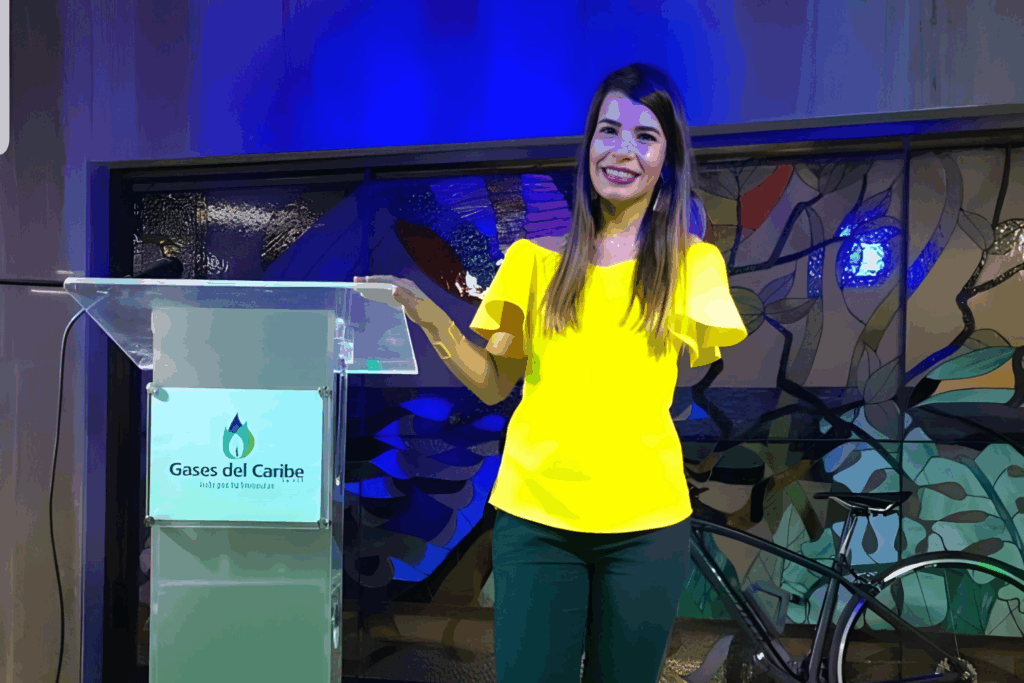 Fotografía de Emily Clavel ofreciendo una conferencia desde un podio con el logotipo de Gases del Caribe, vistiendo blusa amarilla brillante y sonriendo con confianza.