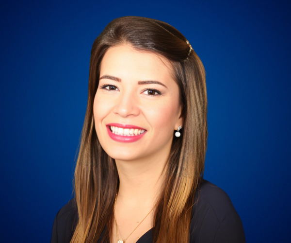 Fotografía de Emily Clavel, sonriendo con una blusa oscura elegante, luciendo aretes de perla, frente a un fondo azul profesional.