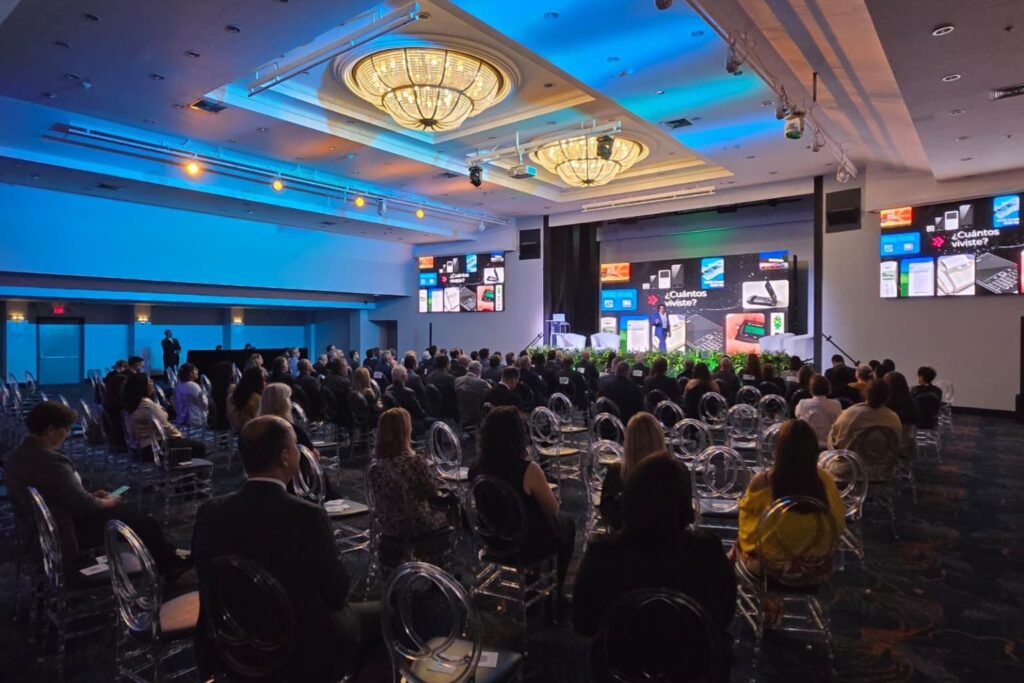 Fotografía de Alejandro Medina impartiendo una conferencia sobre inteligencia artificial en un salón elegante con gran asistencia, acompañado de pantallas digitales que muestran contenido tecnológico.