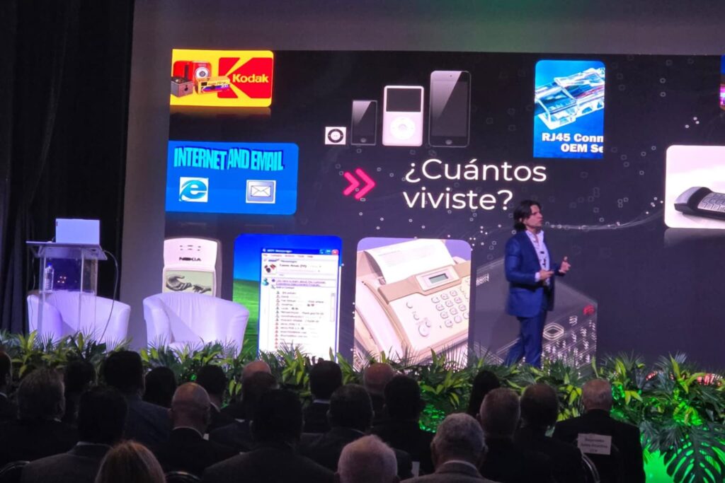 Fotografía de Alejandro Medina dando una conferencia frente a un auditorio lleno, con una pantalla que muestra íconos tecnológicos del pasado como Kodak, Nokia, Internet Explorer y Messenger.
