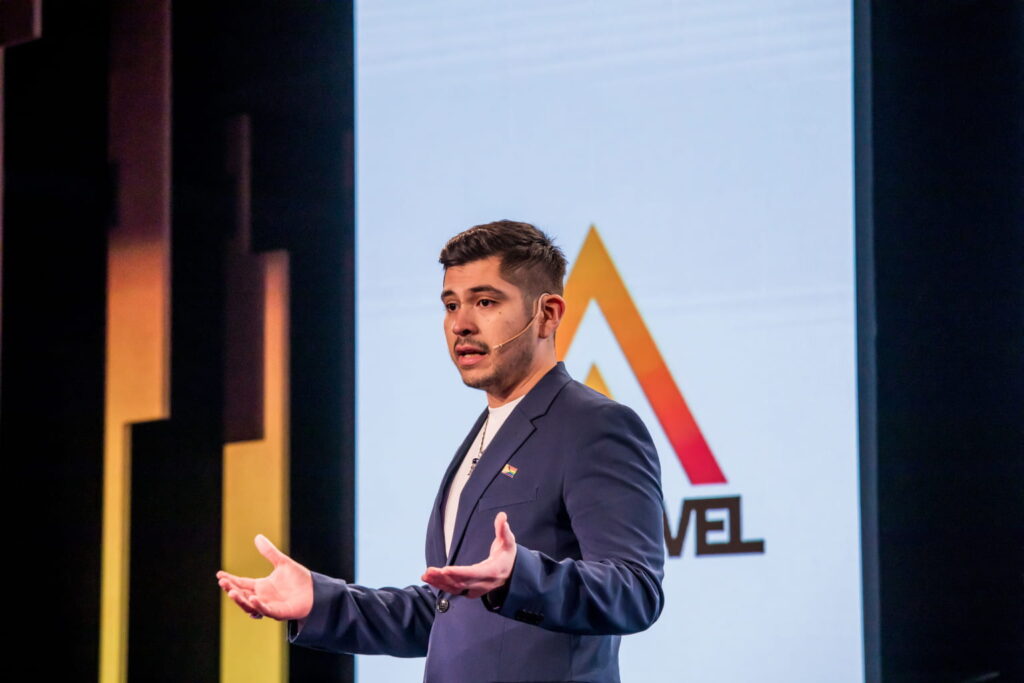 Fotografía de Edgar Dojaque ofreciendo una conferencia en escenario, con micrófono de diadema y fondo con logotipo corporativo, transmitiendo liderazgo y seguridad.