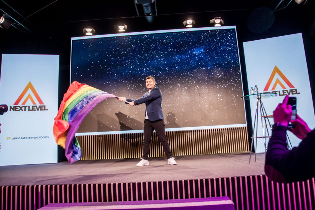 Fotografía de Edgar Dojaque en el escenario de una conferencia Next Level, sonriendo mientras ondea una bandera del orgullo LGBT frente a una pantalla con un fondo estrellado.