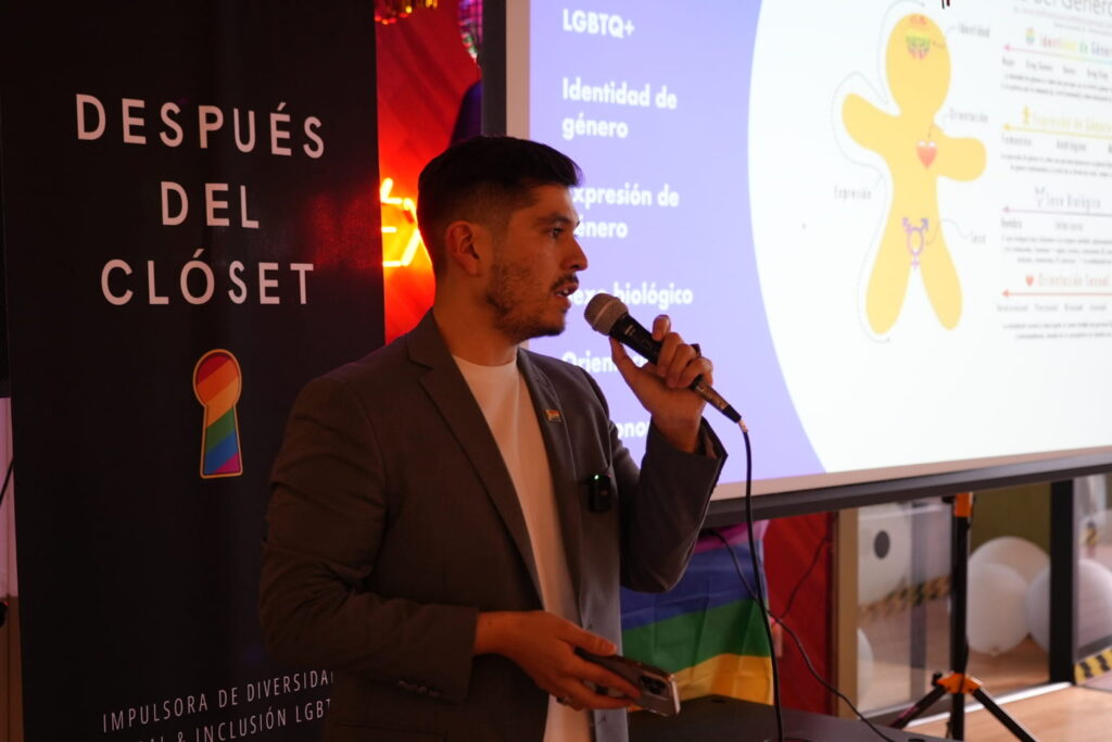 Fotografía de Edgar Dojaque ofreciendo una conferencia sobre diversidad e inclusión LGBTQ+, sosteniendo un micrófono frente a una pantalla con contenido educativo y un banner de “Después del Clóset”.