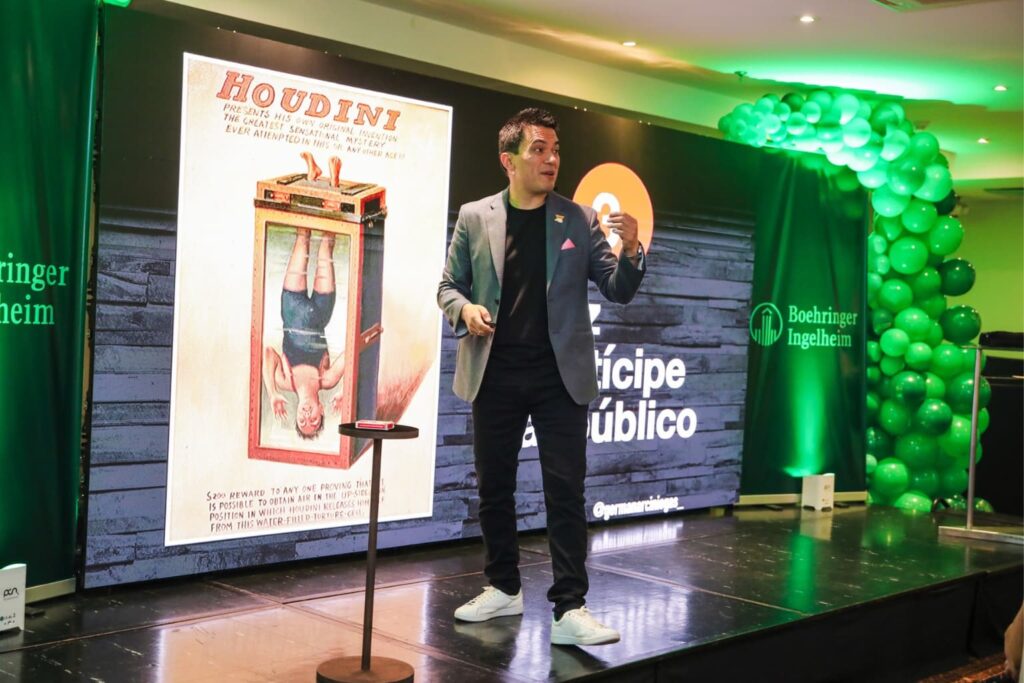 Fotografía de Germán Arciniegas dando una conferencia en evento de Boehringer Ingelheim, con una imagen de Houdini proyectada y decoración con globos verdes.