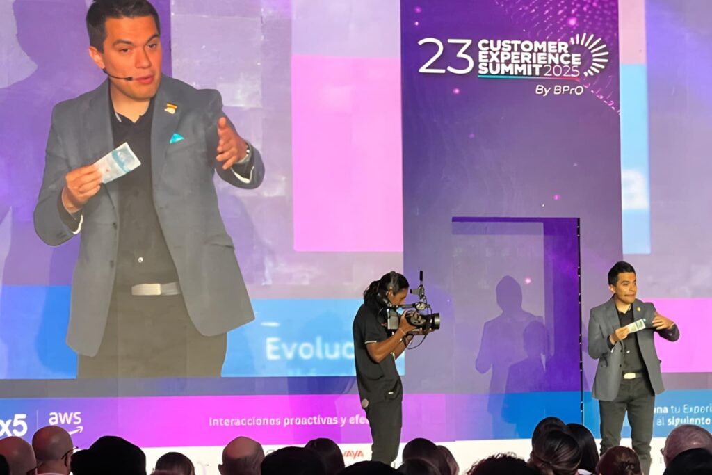 Fotografía de Germán Arciniegas en el escenario del Customer Experience Summit 2025, con micrófono de diadema y sosteniendo un billete mientras es grabado en vivo.