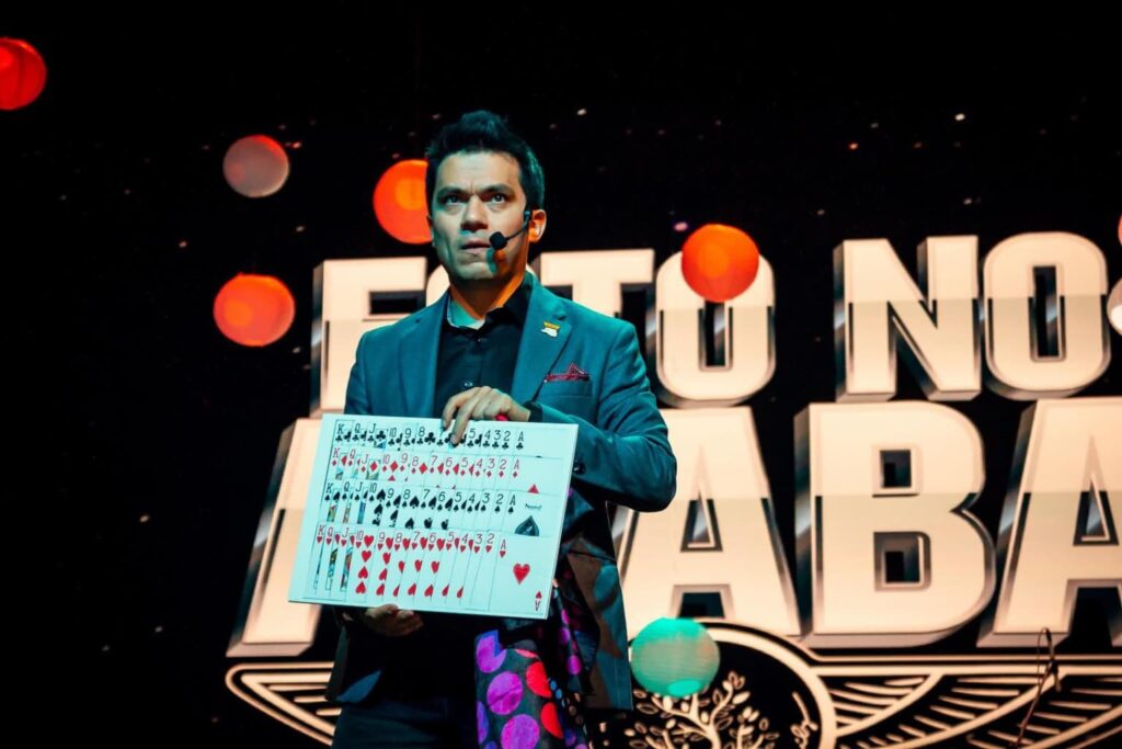 Fotografía de Germán Arciniegas en escenario con micrófono de diadema, mostrando un tablero con cartas de póker frente a fondo con letras grandes y luces decorativas.