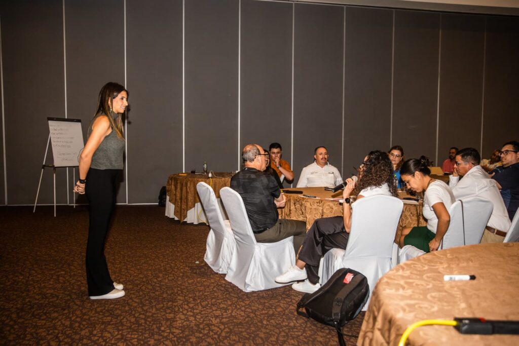 Fotografía de Karem Torres guiando una sesión participativa en salón de conferencias, frente a asistentes sentados en mesas redondas.