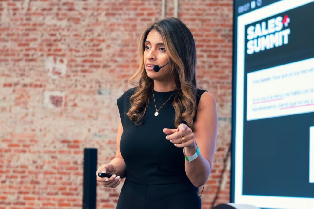 Fotografía de Karem Torres hablando en el escenario del Sales Summit, con micrófono de diadema y fondo de ladrillo expuesto.