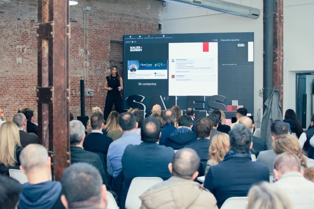 otografía de Karem Torres dando una conferencia en el Sales Summit frente a una audiencia numerosa, con pantalla gigante al fondo y ambiente industrial.