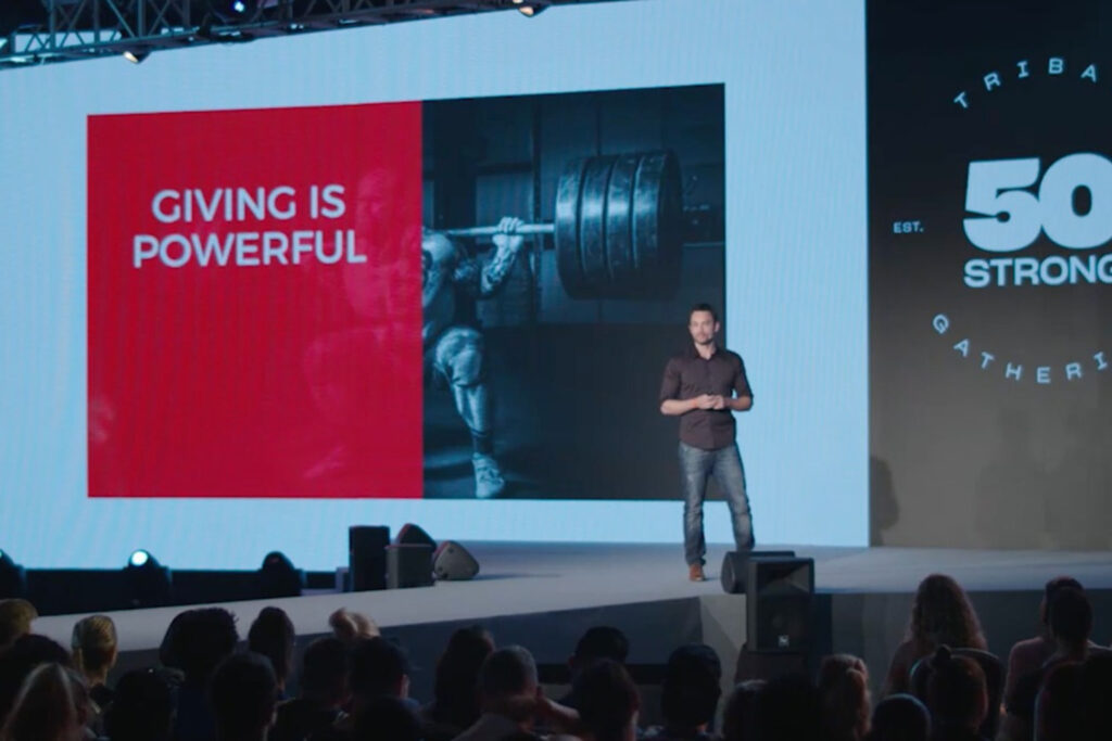 Fotografía de Achi Kushnir en un escenario de conferencia frente a una gran audiencia, con una pantalla proyectando la frase “Giving is Powerful” junto a la imagen de un levantador de pesas, en el evento Tribal Gathering 50 Strong.