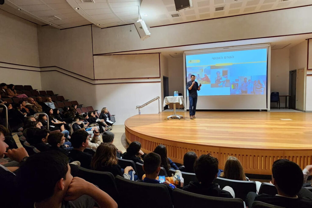 Fotografía de Achi Kushnir ofreciendo una conferencia en un auditorio con público juvenil y adulto, acompañado de una proyección en pantalla con imágenes y texto en hebreo.