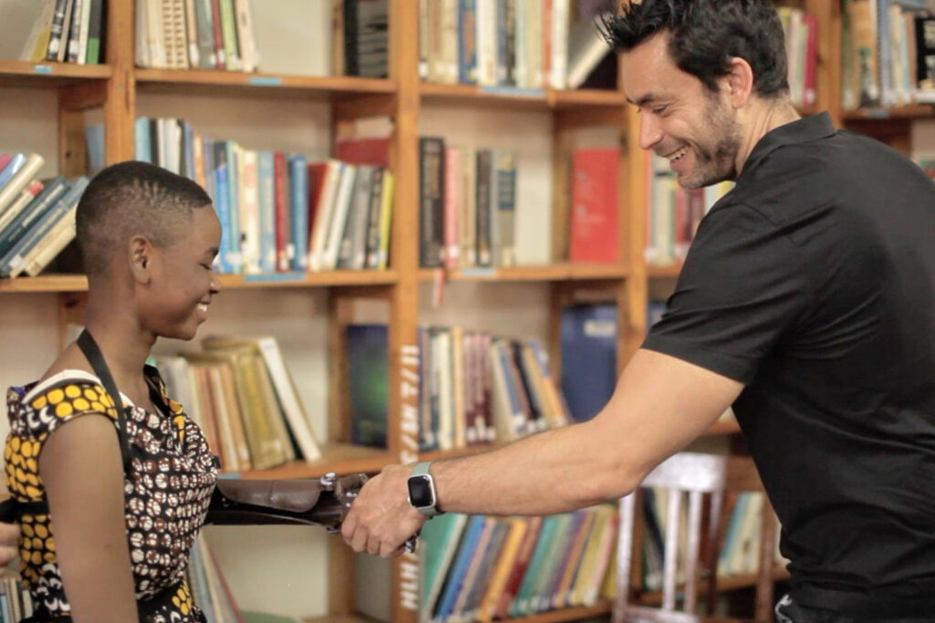 Fotografía de Achi Kushnir sonriendo mientras estrecha la mano de una joven con prótesis de brazo, en una biblioteca con estanterías llenas de libros, transmitiendo inclusión y motivación.