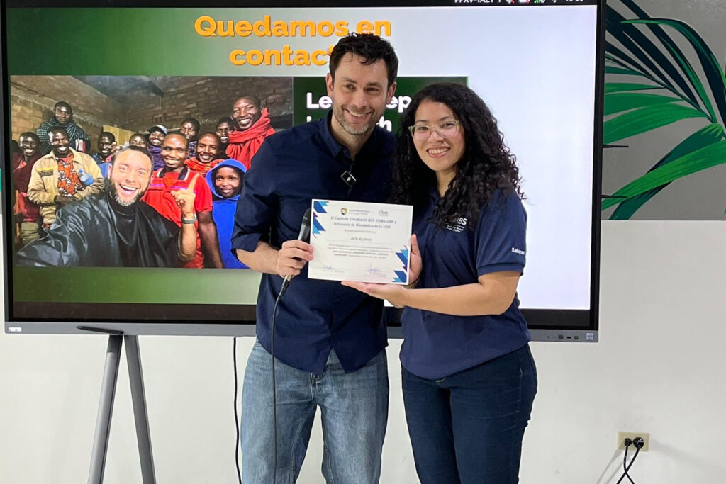 Fotografía de Achi Kushnir recibiendo un reconocimiento junto a una joven en un evento educativo, con una pantalla de fondo que muestra la frase “Quedamos en contacto” y una imagen grupal sonriente.