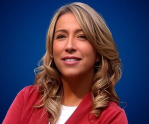 Jennifer Warner sonriendo con fondo azul, vestida con chaqueta roja y cabello rubio ondulado