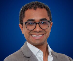 Mauricio Benavides con saco gris, camisa blanca y fondo azul profesional.