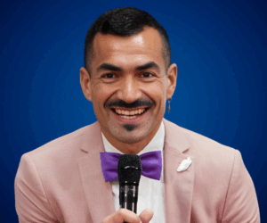 Fotografía de Octavio Díaz Matus sonriendo y sosteniendo un micrófono, vistiendo un traje rosa con moño morado, frente a un fondo azul profesional.