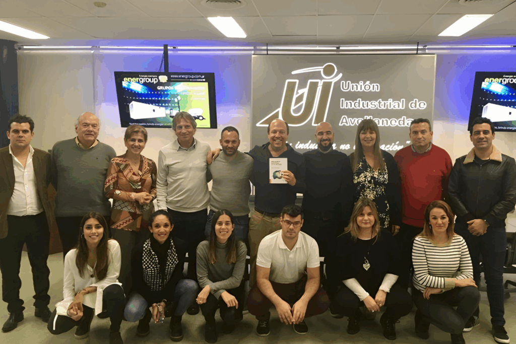 Fotografía de Pablo Nachtigall junto a un grupo de profesionales en un evento realizado en la Unión Industrial de Avellaneda. El conferencista sostiene un libro, al centro del grupo, en un ambiente corporativo con pantallas y logotipos visibles al fondo.
