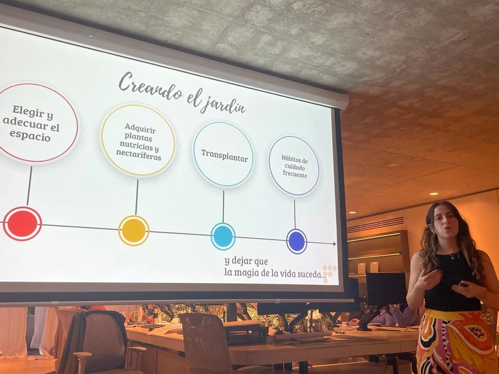 María Belén Sanmauro dando una charla frente a una pantalla con la presentación "Creando el jardín", en una sala de conferencias moderna con iluminación cálida.