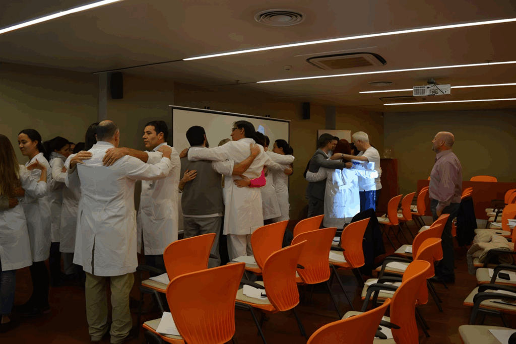 Fotografía de Pablo Nachtigall observando a un grupo de profesionales de la salud abrazándose en una dinámica grupal dentro de un auditorio con sillas naranjas. Los participantes visten guardapolvos blancos, en un entorno iluminado y colaborativo.