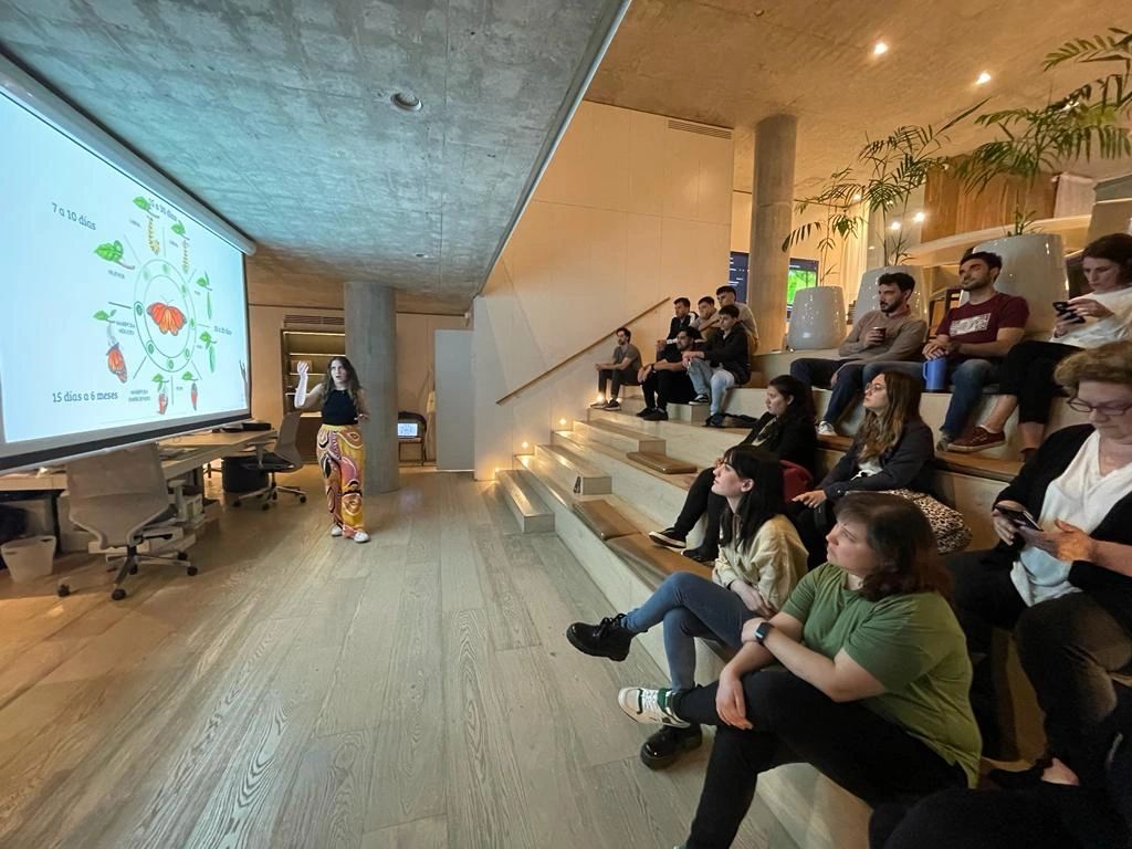 María Belén Sanmauro brindando una charla educativa sobre el ciclo de vida de las mariposas, frente a una audiencia atenta en un auditorio moderno con gradas de madera y ambiente cálido.