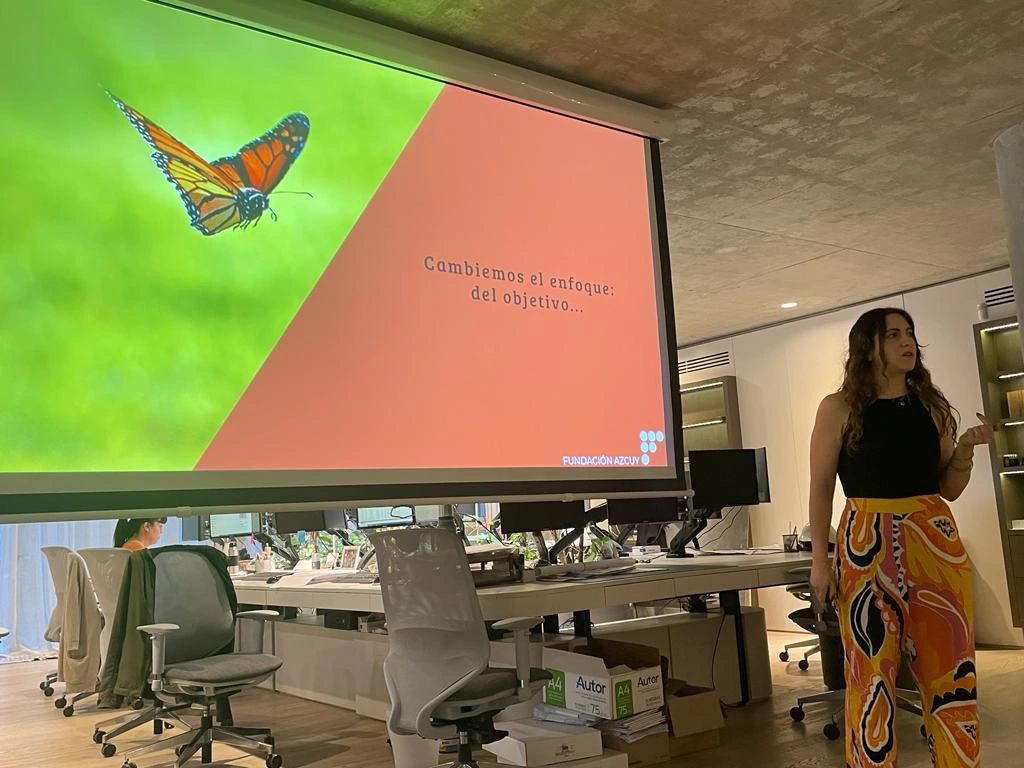 María Belén Sanmauro presentando una diapositiva con una mariposa monarca y la frase “Cambiemos el enfoque: del objetivo…”, en un entorno de oficina moderno.