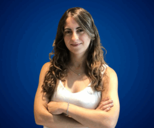 María Belén Sanmauro, con expresión confiada y los brazos cruzados, sobre un fondo azul liso que resalta su presencia profesional.