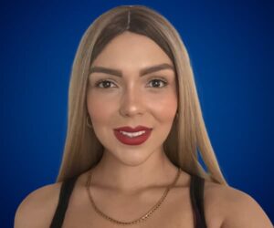 Fotografía de Brenda Caballero con fondo azul, luciendo cabello rubio lacio, maquillaje elegante y cadena dorada.