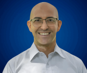 Fotografía de Carlos Bruzzone con gafas y camisa blanca, sonriendo frente a un fondo azul liso con iluminación suave.