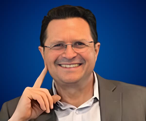 Fotografía de César Camacho, sonriendo con actitud cercana y profesional, vistiendo saco gris y camisa blanca sobre fondo azul sólido.