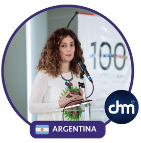 Débora Slotnisky dando una conferencia en Argentina, de pie frente a un micrófono, con fondo institucional y banner de evento "100", luciendo una blusa blanca y collar colorido.