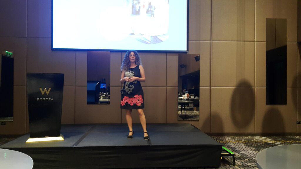 Débora Slotnisky dando una conferencia en el Hotel W de Bogotá, sobre un escenario con pantalla de fondo y vestido negro con detalles florales.