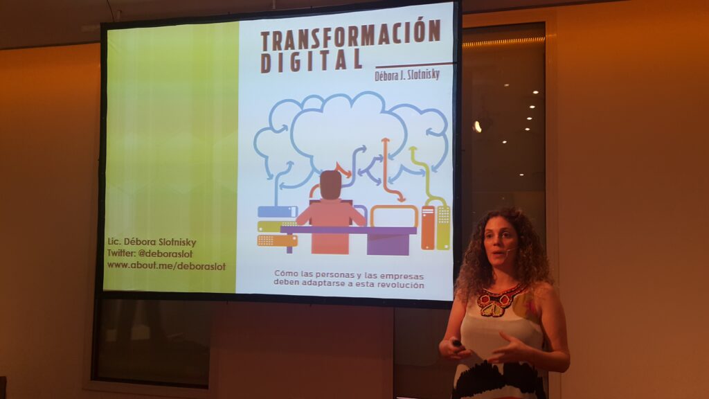 Débora Slotnisky durante una charla sobre transformación digital, frente a una pantalla que muestra la portada de su presentación con ilustraciones tecnológicas y su nombre.