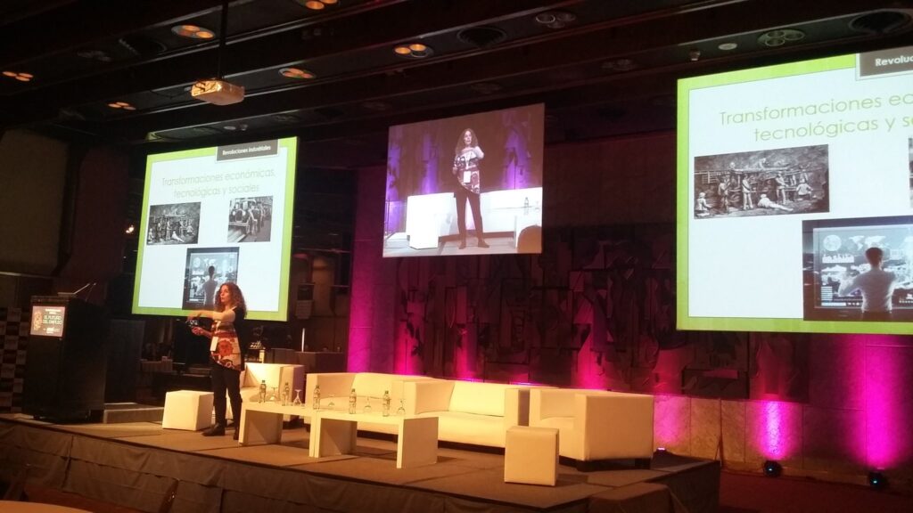 Débora Slotnisky en una conferencia sobre transformaciones económicas, tecnológicas y sociales, hablando desde un gran escenario iluminado con luces violetas, acompañada de una pantalla gigante con imágenes históricas y futuristas.