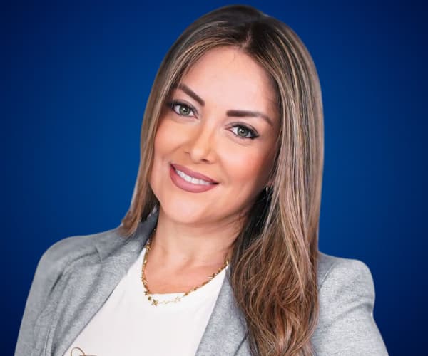 Fotografía de Floribeth Calvo con fondo azul, luciendo una chaqueta gris y una sonrisa profesional en un retrato de primer plano.