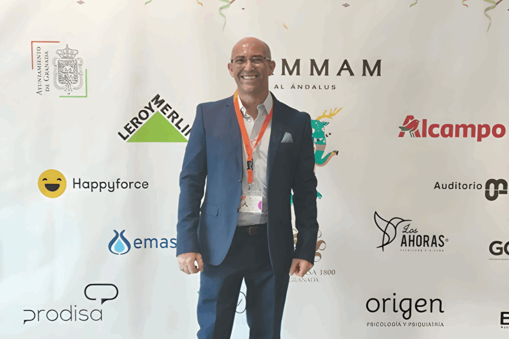 Fotografía de Carlos Bruzzone, conferencista internacional, posando sonriente frente a un mural de patrocinadores durante un evento profesional en Granada.