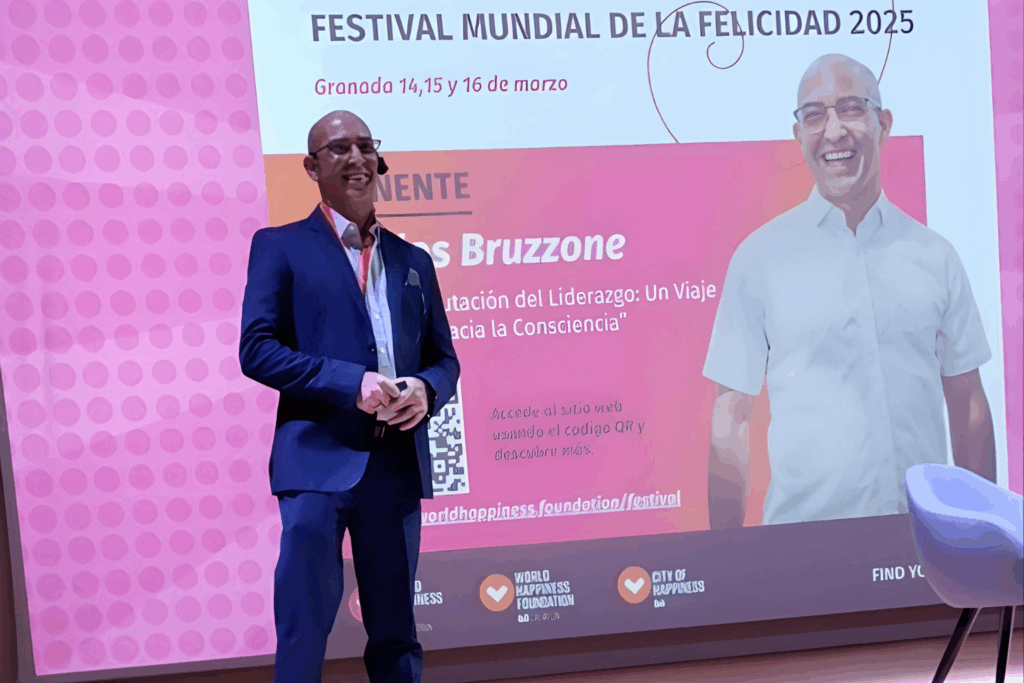 Fotografía de Carlos Bruzzone, conferencista internacional, de pie sobre el escenario del Festival Mundial de la Felicidad 2025 en Granada, con fondo rosa y pantalla de presentación.
