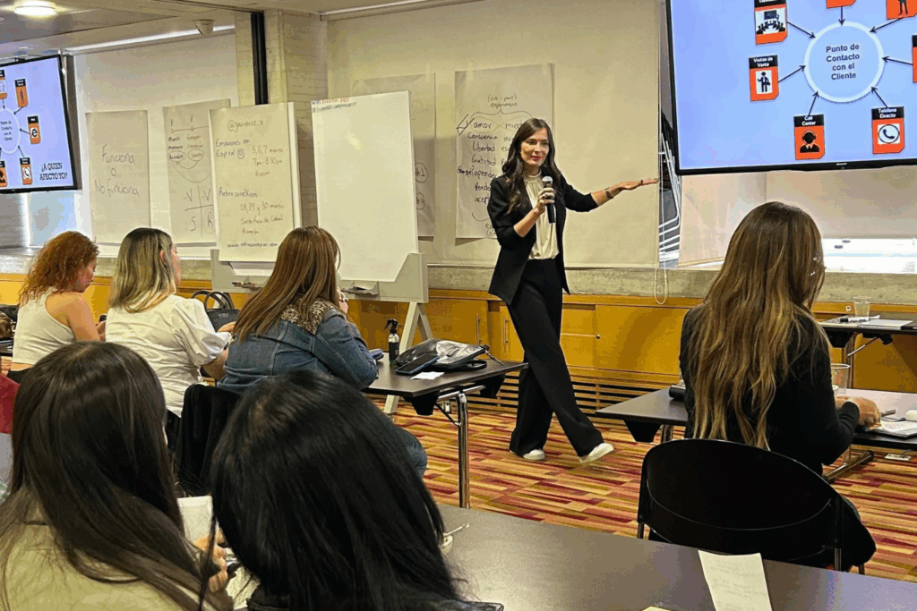 Fotografía de Catalina Serna impartiendo una conferencia en un aula, usando micrófono y presentando sobre puntos de contacto con el cliente, mientras el público toma notas.