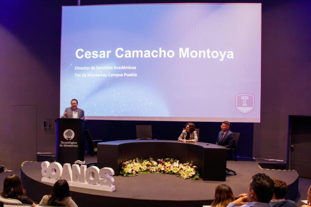 Fotografía de César Camacho Montoya dando una ponencia en el Tecnológico de Monterrey Campus Puebla, acompañado de dos panelistas, en un auditorio decorado por el 80 aniversario de la institución.