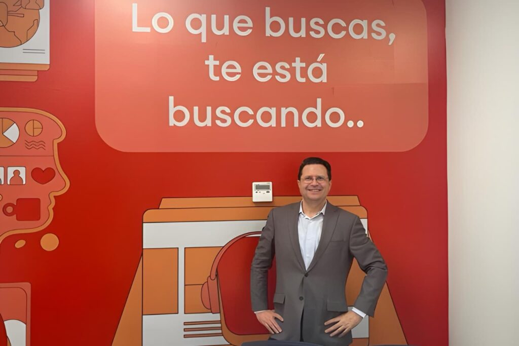 Fotografía de César Camacho Montoya sonriendo frente a un mural rojo con la frase "Lo que buscas, te está buscando", vistiendo saco gris y camisa blanca en un entorno moderno de oficina.