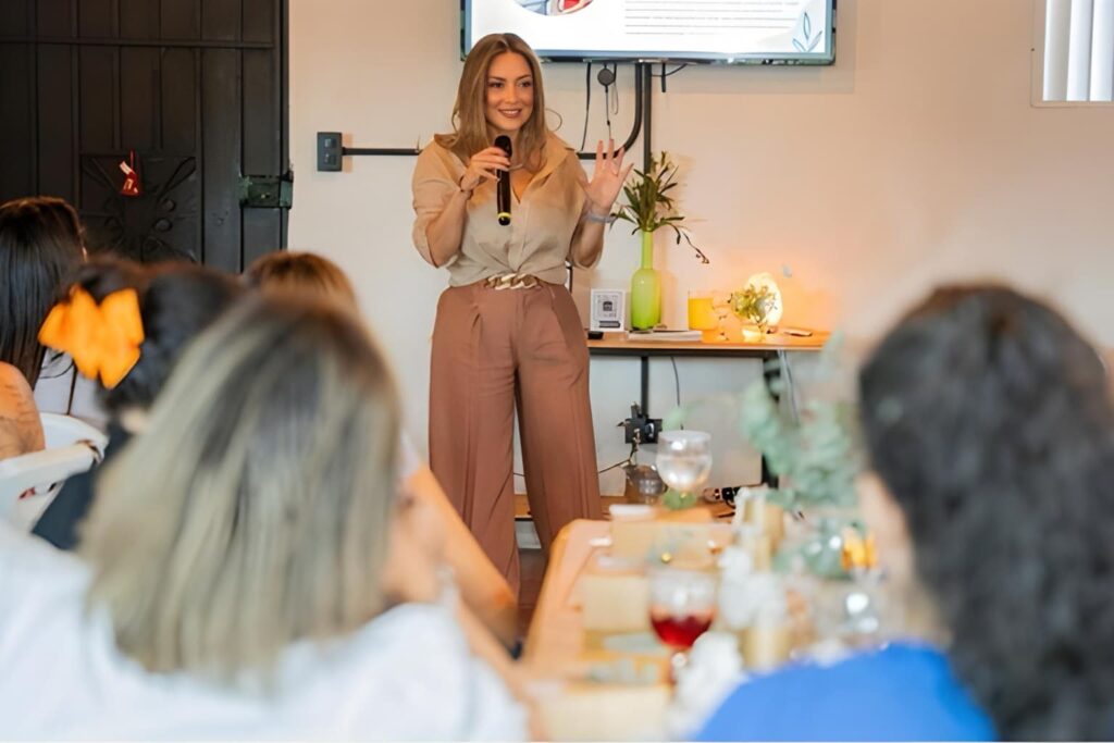 Fotografía de Floribeth Calvo ofreciendo una charla motivacional, sonriendo con micrófono en mano frente a un grupo de asistentes en un ambiente cálido y decorado.