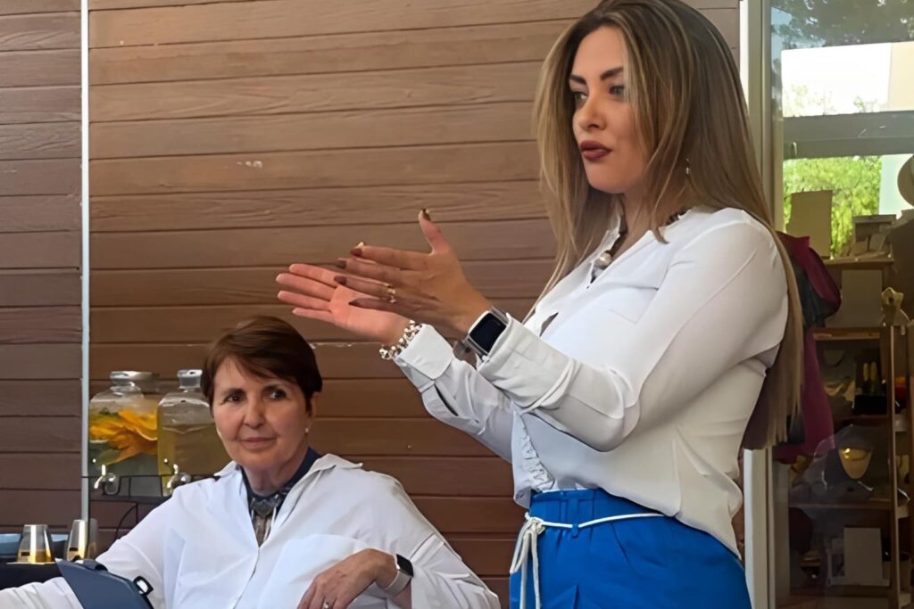 Fotografía de Floribeth Calvo hablando con énfasis durante una conferencia, mientras una asistente escucha atentamente en un entorno acogedor con decoración natural.