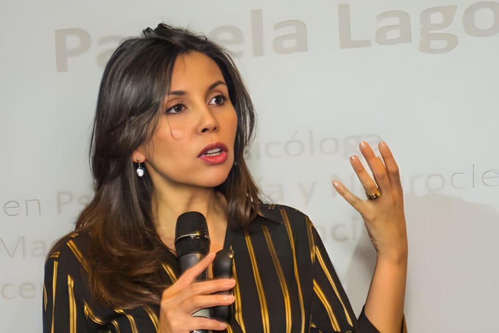 Fotografía de Pamela Lagos, conferencista chilena, hablando apasionadamente con micrófono en mano durante una presentación, frente a un fondo con su nombre proyectado.