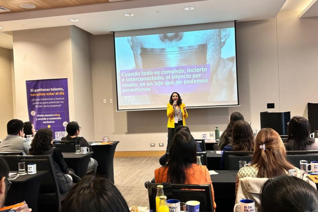 Fotografía de Paola Canessa impartiendo una conferencia, vistiendo chaqueta amarilla y blusa estampada, frente a una audiencia atenta, con una presentación proyectada que destaca la importancia de expresarse en tiempos complejos e interconectados.